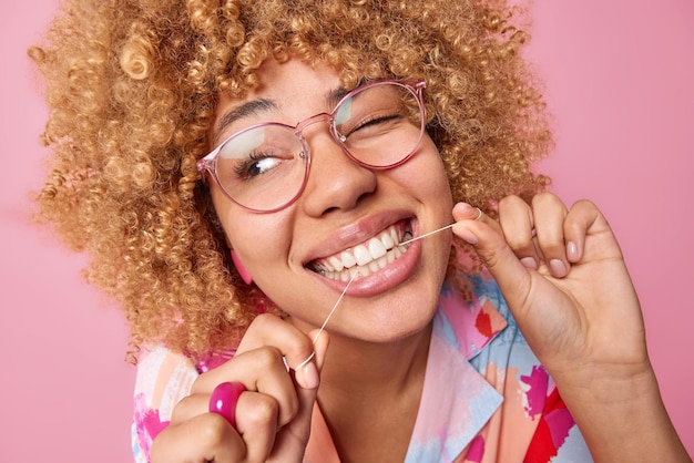 Foto close-up tiro de jovem com cabelos cacheados usa fio dental limpa os dentes usa grandes óculos ópticos pisca o olho e olha de lado isolado sobre fundo rosa sorri amplamente conceito de higiene oral