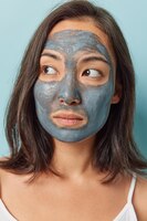Foto close-up tiro de mulher asiática morena pensativa aplica máscara facial de beleza para aumentar a elasticidade das impurezas de purga da pele dos poros poses internas contra fundo azul conceito de bem-estar