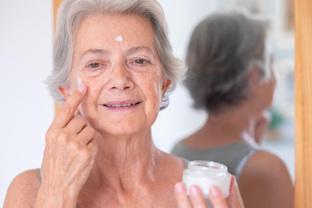 Foto close-up tiro de mulher bonita sênior aplica creme antienvelhecimento no rosto enrugado senhora idosa segurando um produto de frasco cosmético