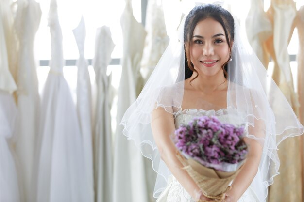 Close-up tiro do buquê de flores do casamento roxo nas mãos da asiática linda noiva em um vestido branco com pé de véu de cabelo seethrough sorrindo olhar para a câmera no fundo desfocado no camarim.