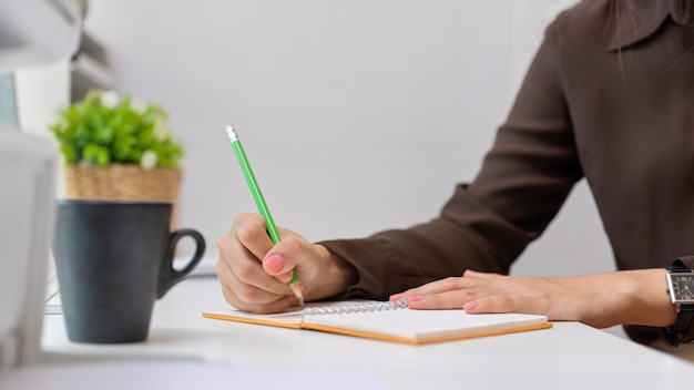 Close-up vista da mão de uma trabalhadora de escritório escrevendo no caderno na mesa branca com o copo e o vaso