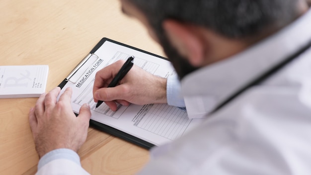 Foto close-up vista das mãos masculinas dos médicos, escrevendo dados do paciente em formulário médico no consultório médico.