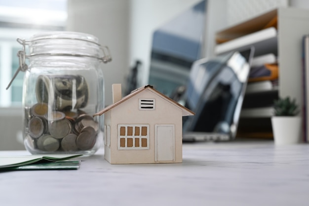 Close-up vista de potes de vidro com moedas e modelo de casa na mesa. planejando economizar dinheiro para comprar uma casa, um imóvel ou um investimento imobiliário.