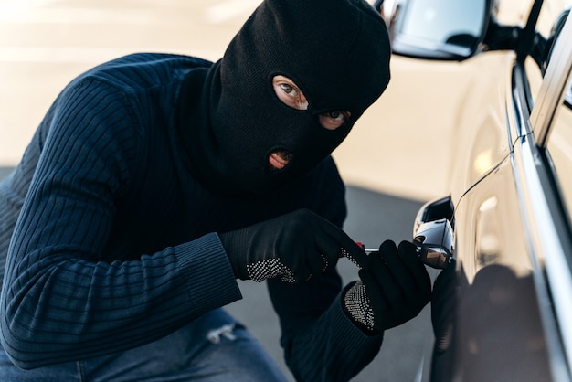 Close-up vista do homem perigoso vestido de preto com uma balaclava na cabeça arromba a fechadura com uma picareta enquanto rouba. Ladrão de carros, conceito de roubo de carros