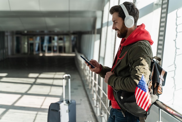 Close-up vista do homem viajante usando fones de ouvido olhando para a tela do smartphone enquanto ouve música e espera seu voo. Cidadãos e conceito de conceito de símbolos americanos