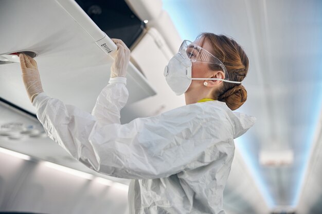 Close-up vista lateral da aeromoça feminina em uniforme protetor branco, olhando para o compartimento da cabine a bordo do avião