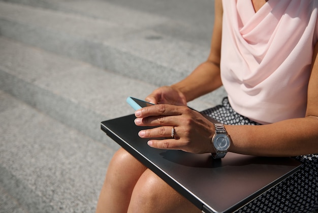 Close-up, vista recortada com foco suave nas mãos de mulher de negócios com relógio elegante, segurando um smartphone, sentado nos degraus com um laptop. Conceito de trabalho empresarial, freelance, online e distante