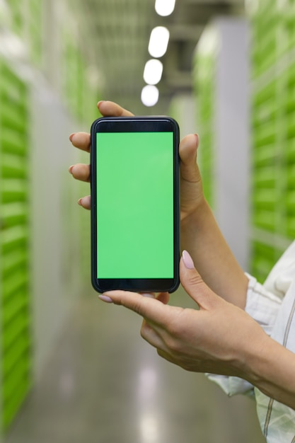 Close vertical de mãos femininas segurando um smartphone com tela verde contra a superfície da instalação de autoarmazenamento