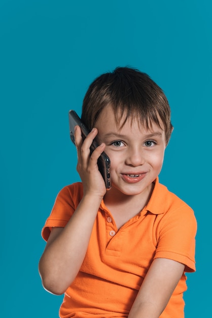 Close vertical de um menino loiro com uma camiseta laranja falando ao telefone sobre um fundo azul