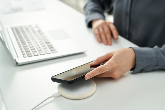 Closeup da jovem empresária com smartphone usando carregador sem fio enquanto trabalhava no laptop em off