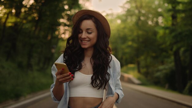 Closeup da linda garota andando ao ar livre Retrato da mulher sorridente mensagem de texto no smartphone no parque ao pôr do sol Pessoa do sexo feminino alegre recebendo boas notícias no celular na estrada