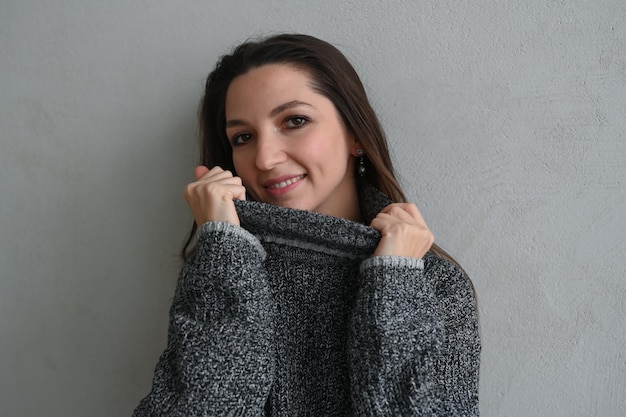 Closeup da linda menina morena puxando o suéter no rosto e sorrindo em pé sobre o espaço de cópia de fundo de concreto