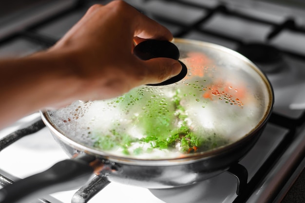 Closeup da mão humana abrindo a frigideira com comida no fogão a gás Foco seletivo na tampa de vidro embaçada da frigideira