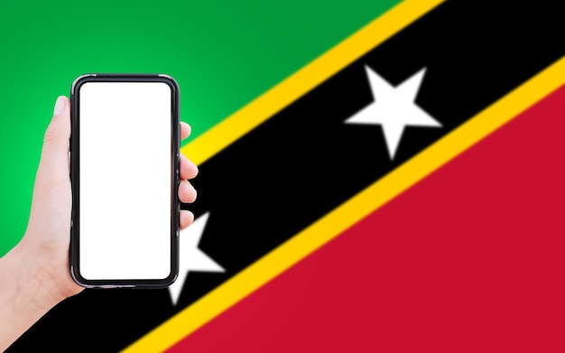 Closeup da mão masculina segurando o smartphone com espaço em branco na tela no fundo da bandeira turva de Kitts e Nevis