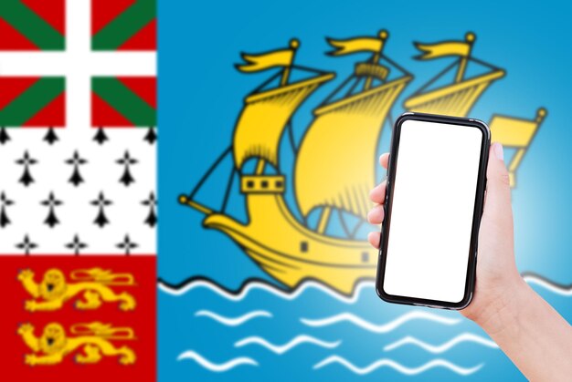 Closeup da mão masculina segurando o smartphone com espaço em branco na tela no fundo da bandeira turva de SaintPierre e Miquelon