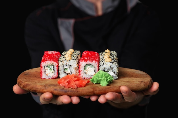 Closeup das mãos de um chef homem em um uniforme preto segurando uma placa de bandeja de madeira cheia de sushi