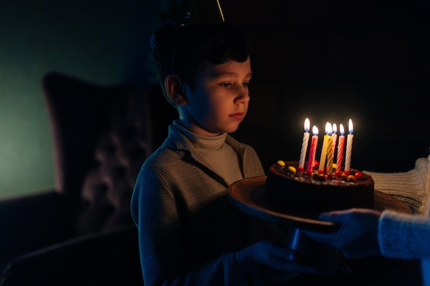Closeup de adorável garotinho fazendo desejo antes de soprar velas no bolo de comemoração de aniversário no quarto escuro