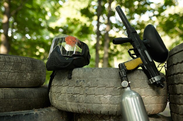 Closeup de arma de paintball e máscara de proteção, ninguém, playground na floresta no fundo. esporte extremo ao ar livre, arma pneumática e balas de tinta, conceito de jogo militar