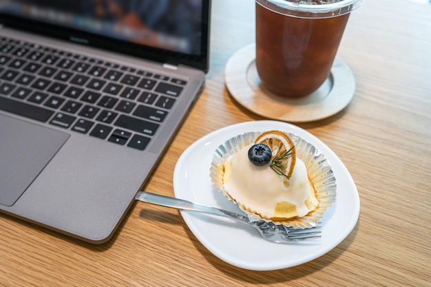 Closeup de café gelado na caneca de xícara e bolo de laranja branco caseiro com teclado laptop na mesa de escritório de madeira na cafeteria no concep de trabalho de negócios de cafedura