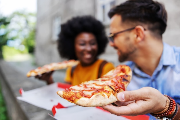 Closeup de casal jovem hippie multicultural sentado ao ar livre e comendo pizza. foco seletivo na mão humana.