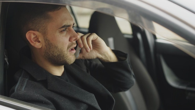 Closeup de empresário estressado xingando e falando ao telefone enquanto está sentado dentro do carro ao ar livre