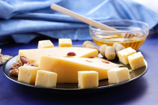 Closeup de fatias de queijo duro com grandes buracos e um guardanapo azul sobre um fundo azul Fatias de queijo amarelo duro com mel de nozes e pãezinhos em um prato preto