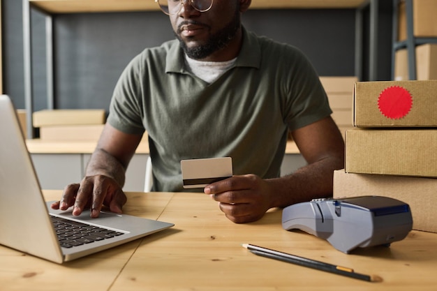 Closeup de homem africano usando laptop para pagar online com cartão de crédito na mesa no armazém