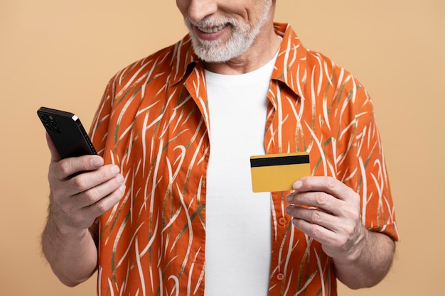 Foto closeup de homem sênior de cabelos grisalhos sorridente, vestindo camisa elegante, segurando o telefone celular e cartão de crédito isolado em fundo bege homem idoso pagando conceito de banco móvel
