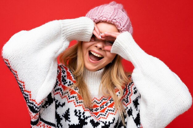 Closeup de jovem positiva feliz linda mulher loira com emoções sinceras, usando chapéu de malha rosa e pulôver de inverno isolado sobre fundo vermelho e mostrando os óculos com as mãos.