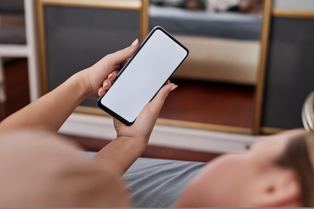Closeup de jovem segurando smartphone com maquete de tela branca enquanto estava deitado na cama e rolando