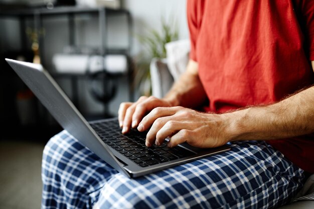 Closeup de jovem sentado na cama com o laptop de joelhos e digitando nele
