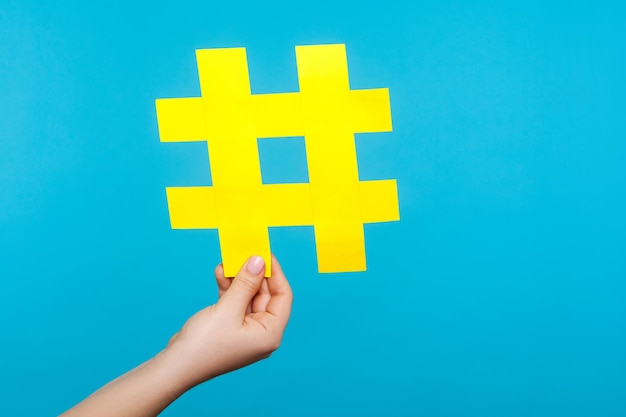 Closeup de mão feminina segurando grande sinal de hashtag de papel amarelo, símbolo de hash de popularidade na internet, tendências de mídia famosas, blogs e conteúdo viral. tiro de estúdio interno isolado em fundo azul