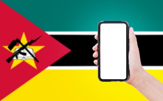 Closeup de mão masculina segurando smartphone com espaço em branco na tela no fundo da bandeira desfocada de Moçambique