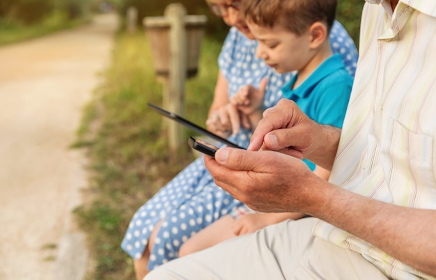 Closeup de mãos de avô tocando um smartphone e neto usando tablet eletrônico com sua avó no fundo. Conceito de valores de geração.