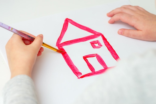 Closeup de mãos de criança desenhando uma casa vermelha com pincel na folha de papel branca. conceito de arte.