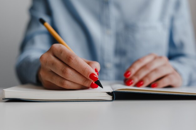 Closeup de mãos de mulher com unhas vermelhas, fazendo anotações em um caderno na mesa.