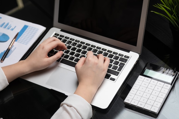 Foto closeup de mãos digitando no laptop com tela preta em branco ao lado de gráficos e calculadora