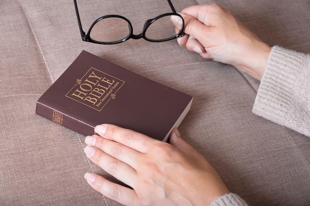 Closeup de mãos femininas com óculos e Bíblia Sagrada no sofá em casa