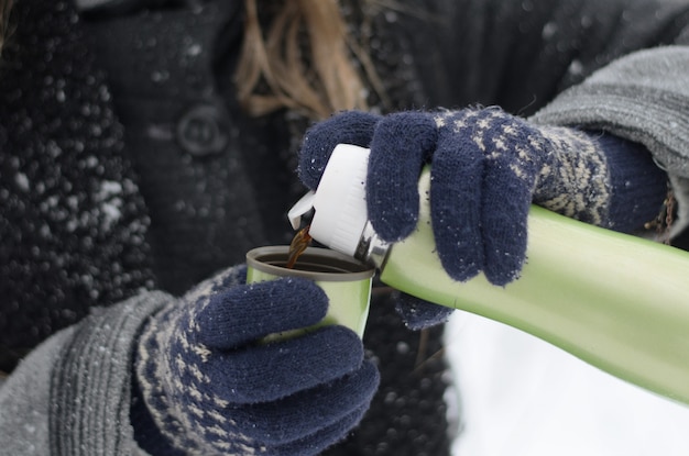Closeup de mãos femininas em luvas de malha, derramando café quente de uma garrafa térmica em um dia frio de neve do inverno
