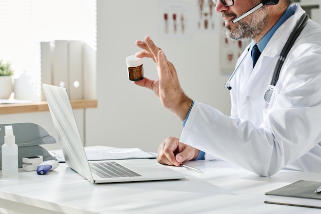 Closeup de médico maduro sentado à mesa em frente ao laptop e apresentando um novo medicamento para sua col