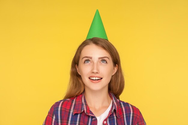 Closeup de menina ruiva sonhadora feliz vestindo cone de festa engraçado na cabeça olhando para cima com expressão sonhadora e fazendo um desejo em sua festa de aniversário estúdio interno tiro isolado em fundo amarelo