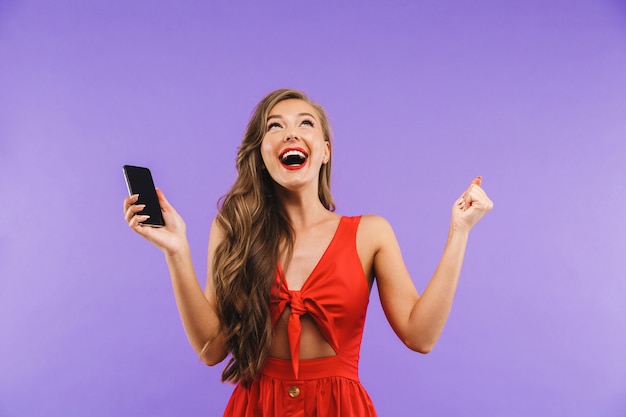 Closeup de mulher bonita feliz usando vestido vermelho, gritando e regozijando-se enquanto segura o smartphone