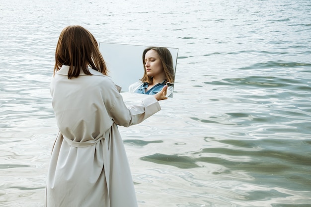 Closeup de mulher em pé em uma costa no vestido azul, segurando um espelho com seu reflexo nele
