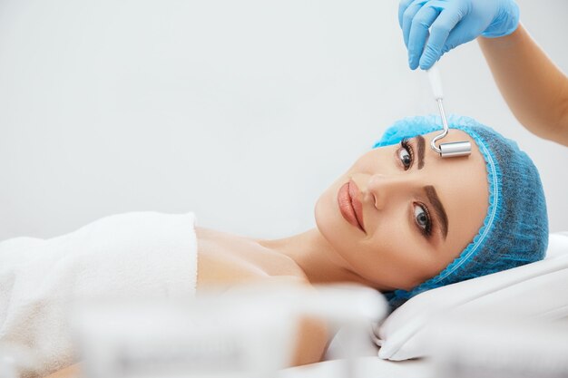 Foto closeup de mulher sorridente com boné azul, deitado no sofá na clínica de cosmetologia e olhando para a câmera. mãos do médico em luvas azuis realizando procedimento com eletrodo para galvanização