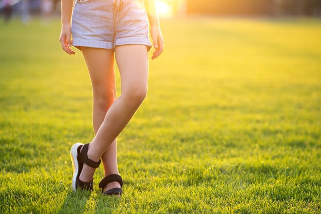 Closeup de pernas de menina criança em shorts jeans, em pé no gramado verde na noite quente de verão.