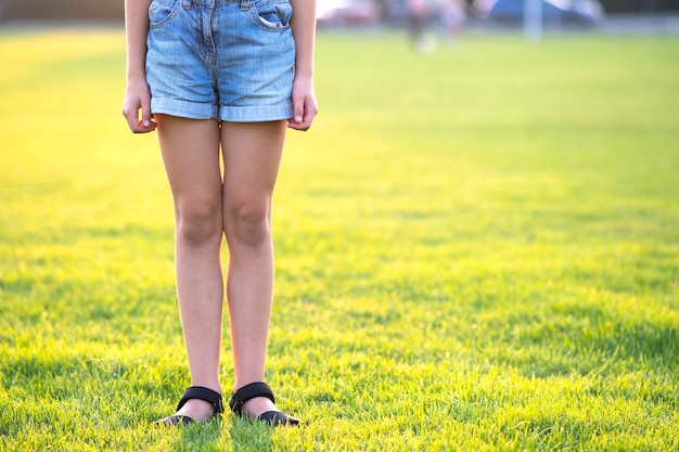 Closeup de pernas de menina criança em shorts jeans, em pé no gramado verde na noite quente de verão.