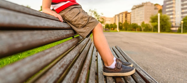 Closeup de pernas de menino com calças curtas sentado no topo de um banco de madeira no parque relaxando em um dia entediado de verão