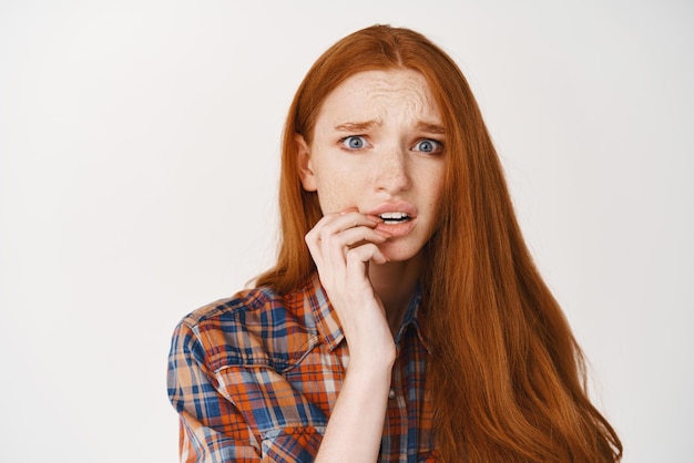 Closeup de uma jovem ansiosa com cabelo ruivo mordendo o dedo e olhando nervoso para a câmera tendo preocupações sobre fundo branco