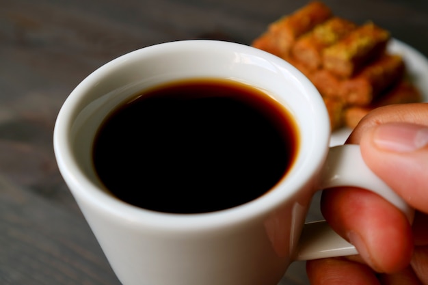 Closeup dedos segurando uma xícara de café turco com bolos de baklava borrados no fundo