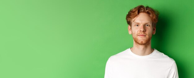 Foto closeup do jovem com cabelo ruivo bagunçado e barba olhando para a câmera em pé sobre fundo verde